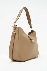 Cartera tipo hobo color beige, de diseño estructurado con cierre superior y una tira vertical con detalle de dije metálico circular en la parte frontal. Cuenta con asa corta trenzada y herrajes dorados.