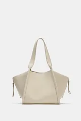 Bolso formato shopper de color beige, con detalle de pespuntes, cremalleras laterales con tiradores, doble asa y compartimento interior extraíble con cierre imantado.