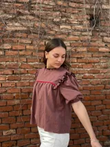 Blusa color chocolate con mangas cortas abullonadas, cuello redondo con volados y detalles de costura en zigzag de color rojo en el pecho y volados oscuros en los hombros.