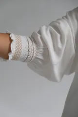 Blusa blanca de corte holgado con cuello redondo fruncido y abertura frontal en V. Presenta mangas tres cuartos con puños elásticos y detalle de tejido texturizado en los mismos.