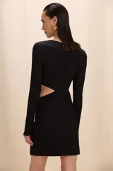 Vestido corto negro de manga larga, con diseño cut-out lateral en la cintura y detalle de aplique metálico dorado en la cadera. Presenta cuello redondo y silueta ajustada.