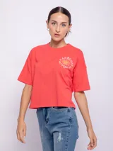 Remera corta de manga corta color coral o rojo anaranjado, con cuello redondo y un estampado circular en el pecho que incluye un sol sonriente y el texto "HARDCORE HAPPINESS".