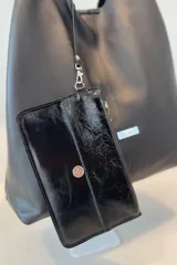 Bolso tipo tote de gamuza en color verde oliva, con diseño minimalista y asa integrada. Presenta una placa metálica con el logo de la marca en la parte inferior central y detalles de textura en el asa.