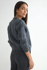 Cardigan de punto trenzado color crema con cuello redondo y cierre frontal de botones grandes en tono dorado. Presenta mangas largas con puños acanalados y un diseño de tejido con textura.