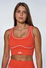 Conjunto deportivo de dos piezas color naranja, compuesto por un top de escote redondo con costuras a la vista en contraste y un short de tiro alto con costuras a la vista. El top presenta el logo de la marca estampado en el frente.