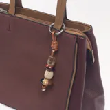 Cartera de mano color bordó con paneles laterales texturizados, manijas superiores en color tostado y dije colgante con cuentas decorativas. Incluye correa larga ajustable y extraíble.