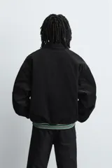 Cazadora estilo varsity confeccionada en pana de algodón azul, con interior ligeramente acolchado. Presenta cuello solapa, cierre frontal con botones a presión, bolsillos laterales de vivo y acabados en rib a rayas en puños y cintura. Incluye múltiples parches bordados decorativos en el frente.