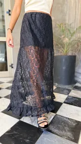 Falda larga de encaje negro con diseño de capas, presentando una sección superior opaca y una sección inferior transparente con terminación de volado en el ruedo.