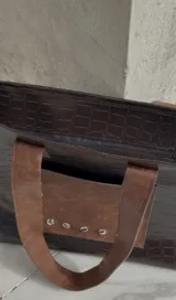 Bolso tote negro con textura símil cuero de cocodrilo y bolsillo frontal marrón con tachas plateadas. Tiene doble asa de hombro marrón y cierre superior.