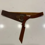 Cinto ancho de cuero marrón con diseño asimétrico, que presenta un aro metálico central y una tira de gamuza para anudar.