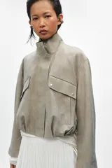 Chaqueta corta de ante con cuello alto, manga larga y cierre frontal con cremallera y botones a presión. Presenta bolsillos frontales con solapa y bajo abullonado con pliegues.