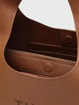 Cartera tipo hobo Marc Jacobs modelo The Sack, confeccionada en cuero granulado color marrón. Presenta un diseño minimalista con el nombre de la marca y el modelo estampados en el frente y una correa integrada al cuerpo de la cartera.