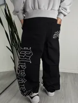 Pantalón baggy negro de corte holgado con bordados en hilo blanco. Presenta un diseño tipográfico estilo gótico en la pierna derecha y un detalle gráfico en el bolsillo trasero.