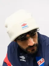 Gorro tipo beanie de tejido de punto color blanco crema, con parche frontal que incluye el logo de Umbro y la inscripción Nacional.