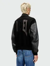 Campera estilo universitaria (varsity jacket) negra con mangas de cuero sintético negro y cuerpo de lana negra. Presenta un parche bordado grande en el pecho y cierre frontal de botones a presión. Tiene cuello, puños y cintura acanalados.