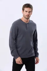 Remera de algodón texturada color gris oscuro, de manga larga, con cuello redondo y botones en la parte frontal. Presenta un pequeño logo bordado en el pecho y un parche decorativo en la manga izquierda.