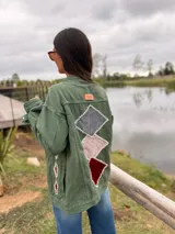 Campera de jean color verde oliva, con diseño de figuras geométricas en terciopelo aplicadas en la espalda y mangas, bordadas a mano con costuras decorativas. Presenta un corte holgado y etiqueta de cuero en la parte superior de la espalda.