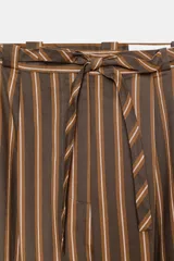 Pantalón de tiro medio con diseño tipo ballon, estampado a rayas verticales en tonos marrones y blancos. Presenta pliegues frontales, bolsillos laterales ocultos y cinturón del mismo tejido integrado en la cintura.