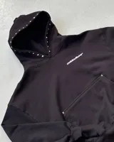 Canguro negro de corte holgado con capucha, decorado con pequeñas tachas metálicas en el borde de la capucha y en el bolsillo frontal. Presenta un estampado pequeño con el logo de la marca en el pecho.