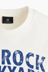 Remera de algodón blanca con estampado azul estilo universitario de toro y texto "Red Rock Stockyard". Presenta cuello redondo, mangas cortas y hombros caídos.