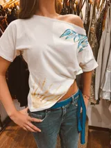 Remera de algodón color blanco con escote asimétrico de hombro descubierto. Presenta un diseño pintado a mano con un sol en la parte inferior y alas en la parte superior.