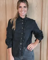 Camisa de denim negro con efecto lavado, cuello alto con volados, cierre frontal con botones y mangas largas con puños elásticos fruncidos.