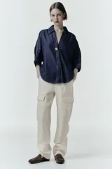 Camisa de denim azul oscuro con corte amplio, cuello solapa, escote en pico y mangas tres cuartos. Presenta un aplique metálico dorado en el centro y bajo asimétrico.