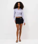 Short pollera mini de tiro bajo, confeccionado en bengalina negra. Presenta un diseño de cintura cruzada con detalle de hebilla ovalada metálica en el frente.