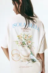 Remera oversized de punto suave de algodón color beige, con cuello redondo acanalado, hombros caídos y diseño estampado en el pecho con la inscripción "Taormina Sicily" en color azul.