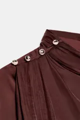 Blusa de tejido satinado color bordó, con cuello subido drapeado y manga larga con puños. Presenta detalle de pliegues frontales y cierre con botones laterales en el cuello.