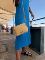 Bandolera pequeña de diseño rectangular en color beige, confeccionada en cuero sintético. Presenta una correa larga ajustable del mismo material y cierre superior con cremallera. Incluye el logo de la marca grabado en la parte frontal.