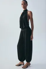 Mono largo negro con cuello halter, hombros descubiertos, parte superior drapeada y piernas anchas con bajo acabado en puño elástico.