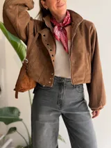 Chaqueta corta de efecto gamuza con cierre frontal y botones. Presenta mangas tipo globo con puños elásticos y detalle de aros metálicos decorativos en la solapa. Cuenta con ajuste lateral en la cintura y forro interior.