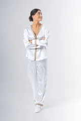 Pijama blanco de satén elastizado, compuesto por una camisa de manga larga con escote en V, abotonada adelante y con puntilla en escote y puños, y un pantalón recto con bolsillos laterales, elástico en la cintura y puntilla en el ruedo.