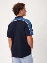 Remera polo gris melange de ajuste holgado, con cuello y cierre frontal de botones. Presenta detalles en azul marino y blanco en los hombros y mangas, y el logo de la marca bordado en el pecho.