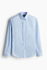 Camisa de tela azul claro con tratamiento de planchado fácil, cuello inglés, tapeta clásica y canesú en la espalda. Mangas largas con tapeta y botón, y cierre ajustable de botón en los puños. Bajo levemente redondeado. Corte ajustado que realza el contorno del cuerpo.