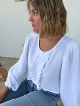 Blusa blanca de algodón con textura arrugada, cuello redondo, mangas largas abullonadas con puños elásticos y cierre frontal con botones y volantes.
