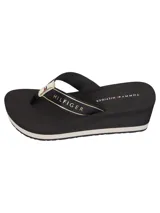 Ojotas de mujer con plataforma tipo cuña de color negro. Las tiras son de textil negro con una banda central que lleva el texto "HILFIGER" en dorado y el logo de Tommy Hilfiger en la unión con la base.