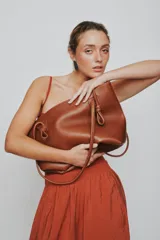 Cartera tote de cuero suave color marrón, con diseño amplio y líneas simples, con tiras de hombro decoradas con nudos artesanales y pequeño logo grabado en el centro.