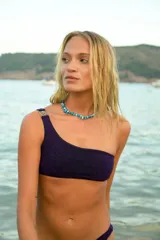 Bikini de tela lúrex color violeta con acabado brillante. El corpiño es tipo top de un solo hombro con hebilla metálica decorativa en el tirante. La bombacha es de corte vedetina.