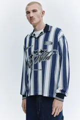 Camiseta de fútbol de manga larga confeccionada en malla estampada, con diseño de rayas verticales en azul marino y blanco. Presenta cuello polo negro con ribete blanco, cierre frontal parcial en V, y puños elásticos negros con líneas blancas. Cuenta con estampado frontal con gráficos de estrellas y la inscripción "Run Wild".