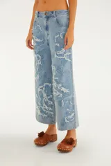 Pantalón jean de tiro medio y corte recto, color azul claro, con grandes bordados florales en tono sobre tono y detalles deshilachados. Cuenta con bolsillos funcionales y cierre frontal con botón y cremallera.