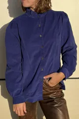 Camisa de pana azul con cuello alto tipo mao con volados, cierre frontal de botones y mangas largas con hombros ligeramente abullonados.