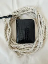 Billetera pequeña de cuero negro con textura de cocodrilo. Cuenta con cierre metálico perimetral y una correa de muñeca extraíble. Incluye compartimentos internos para billetes y un bolsillo con cierre para monedas.