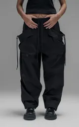 Pantalón de estilo utilitario y corte oversize, confeccionado en textil técnico liviano. Presenta cintura elástica con cordón ajustable, bolsillos laterales envolventes de gran tamaño con cordones y tancas de ajuste, y elásticos regulables en los tobillos.
