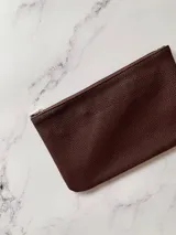 Sobre billetero de cuero sintético con textura granulada en color borra de vino. Cuenta con cierre superior y bolsillo interno.