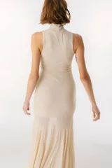 Vestido largo de color beige, con cuello alto y diseño sin mangas. Presenta un corte ajustado en la parte superior y una falda con paneles de tela semitransparente que aportan movimiento y fluidez.