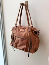 Bolso maternal de cuero marrón con doble asa de hombro, correa ajustable, múltiples bolsillos frontales y laterales con cierre, y detalles de tiras trenzadas.