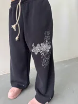 Pantalón de jogging negro de corte baggy, confeccionado en algodón. Presenta una estampa plastisol con bordado decorativo en la pierna izquierda, cintura elástica con cordón largo ajustable y botones en el ruedo para regular la botamanga.
