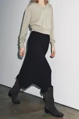 Jersey de punto color beige claro, con cuello en V y manga larga. Presenta detalles de botones decorativos en los hombros y en el ruedo inferior, sugiriendo un diseño desmontable o con aberturas ajustables.